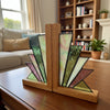 A pair of Art Deco style stained glass bookends on a light marble countertop with a blurred dining room background. The bookends feature geometric fan-shaped stained glass panels in mint green, pale pink, and lavender with black lead lines, supported by a warm oak wood base and central upright.