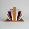 A stained glass bookend with geometric fan-shaped panels in burgundy, white, and lavender, held together by two vertical oak wood posts on a matching oak base, set on a light marble countertop.