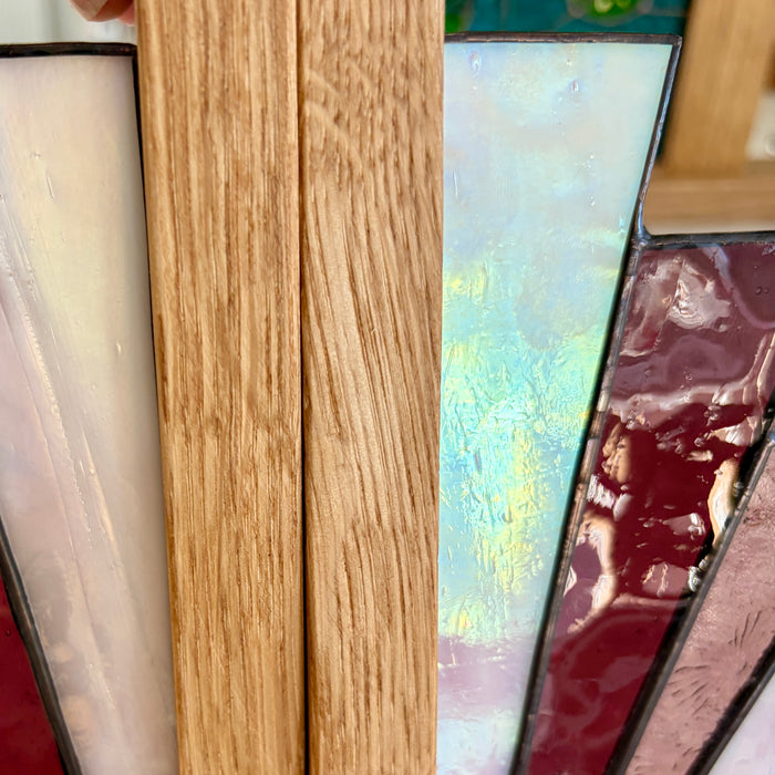 Close-up of stained glass panels with a prominent light oak wood frame in the center, flanked by cream, iridescent blue-green, and deep burgundy glass sections.