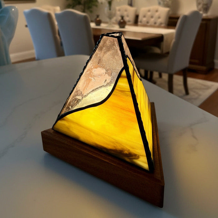 A Tiffany-style stained glass table lamp with a geometric pyramid design, featuring a warm yellow and orange glass panel, black lead lines, and a dark wood base, shown on a dining table in a beautifully lit dining room.