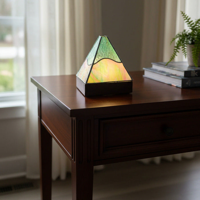 A geometric stained glass lamp with a triangular pyramid design, featuring textured green and iridescent yellow glass panels held together by dark metal frames, shown lit on a gray tabletop.