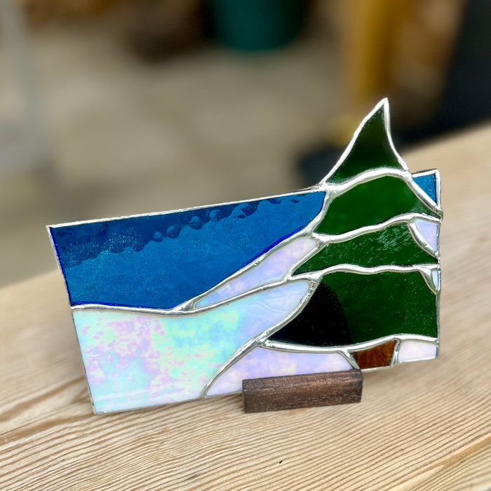 A stained glass ornament depicting a winter landscape with pine trees, set on a small wooden stand and photographed on a light wood surface.