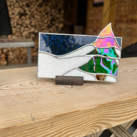 A stained glass window panel depicting a stylized female figure with colorful abstract hair, resting on a wooden surface in a workshop setting.