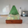 A triangular mountain-shaped decorative object with a textured green top section, iridescent multicolor lower section, and a medium-brown wooden base, photographed on a light marble-like surface with other similar objects blurred in the background.