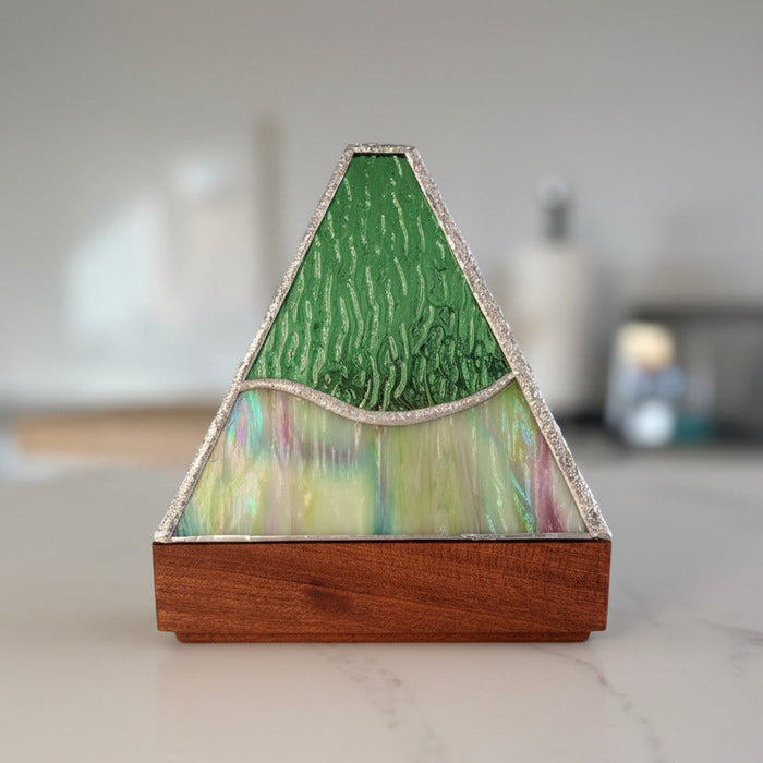 A triangular mountain-shaped decorative object with a textured green top section, iridescent multicolor lower section, and a medium-brown wooden base, photographed on a light marble-like surface with other similar objects blurred in the background.