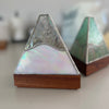 Two triangular decorative boxes with iridescent abalone shell panels, silver-toned metal borders, and wooden bases, shown on a light tabletop with a softly blurred background.