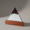 A triangular pyramid-shaped decorative object with an iridescent mother-of-pearl front panel, a dark reddish-brown top section, and a silver metal frame, displayed on a wooden base on a light tabletop. Other similar items are blurred in the background.