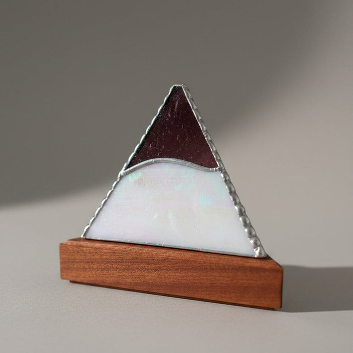 A triangular pyramid-shaped decorative object with an iridescent mother-of-pearl front panel, a dark reddish-brown top section, and a silver metal frame, displayed on a wooden base on a light tabletop. Other similar items are blurred in the background.