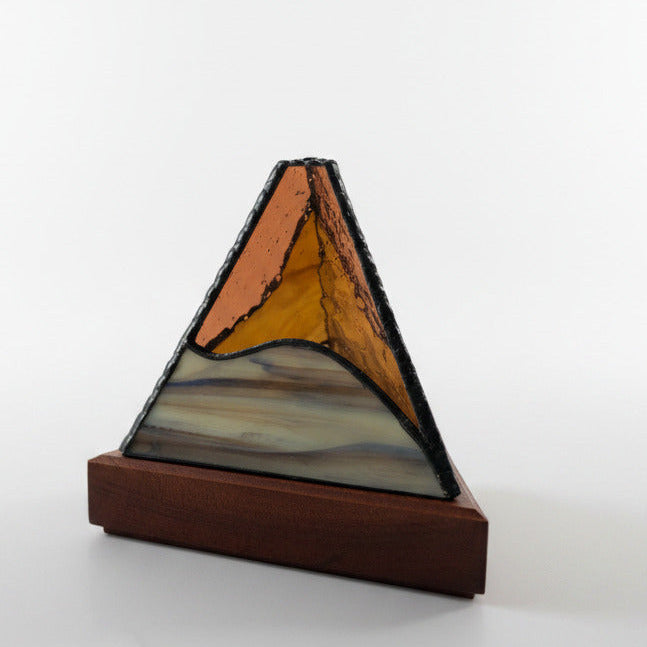 A triangular stained glass pyramid with a copper-gold top section, amber center, and marbled blue, white, and gray lower section, mounted on a polished wooden base. Other similar glass pyramids are blurred in the background on a light marble surface.