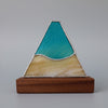 A triangular stained glass window piece depicting a mountain landscape, featuring textured teal blue and green glass on the upper half and a pale yellow glass section on the lower half, mounted on a wooden base.