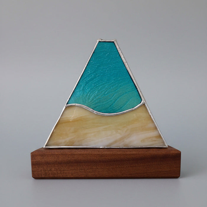 A triangular stained glass window piece depicting a mountain landscape, featuring textured teal blue and green glass on the upper half and a pale yellow glass section on the lower half, mounted on a wooden base.