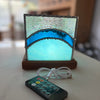 A square LED light box with a stained-glass-like design featuring a textured silver top, a bright blue wave pattern, and a glowing light blue center, sitting on a wooden base. A white USB charging cable and a black remote control are placed in front on a light-colored tabletop.