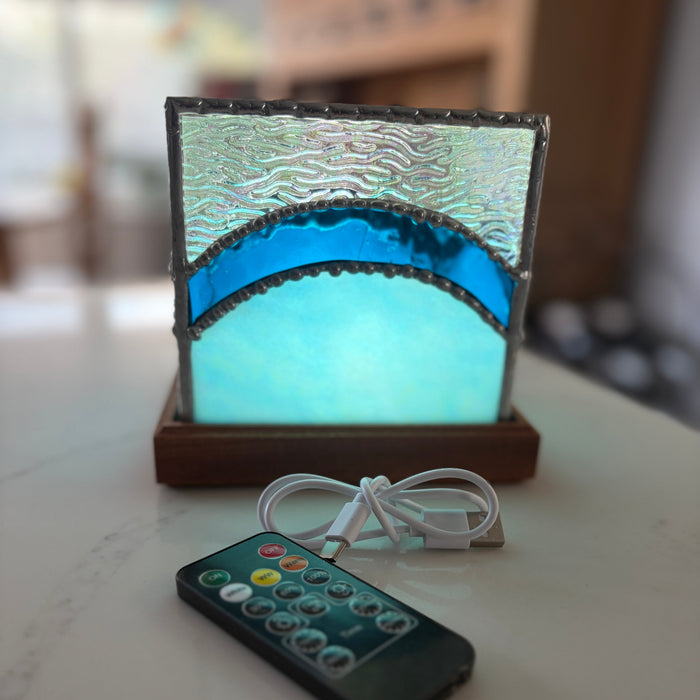 A square LED light box with a stained-glass-like design featuring a textured silver top, a bright blue wave pattern, and a glowing light blue center, sitting on a wooden base. A white USB charging cable and a black remote control are placed in front on a light-colored tabletop.