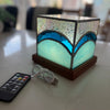 A square LED mosaic lamp with a blue and light green wave design, resting on a wooden base next to a remote control and USB cable on a light-colored table.