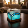 A square decorative lamp or box with a wooden base, featuring textured silver glass on top and a glowing light blue glass front, set on a light-colored tabletop with a blurred indoor background.