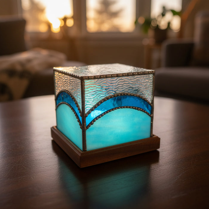 A square decorative lamp or box with a wooden base, featuring textured silver glass on top and a glowing light blue glass front, set on a light-colored tabletop with a blurred indoor background.