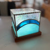 A square decorative lamp with a glowing aqua blue glass body, a textured iridescent silver top panel, and a dark blue metallic arched band, set on a dark wood base.