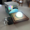 A square LED night light with a glowing white globe on a wooden base, shown with its textured metallic box packaging and a black remote control on a light marble tabletop.