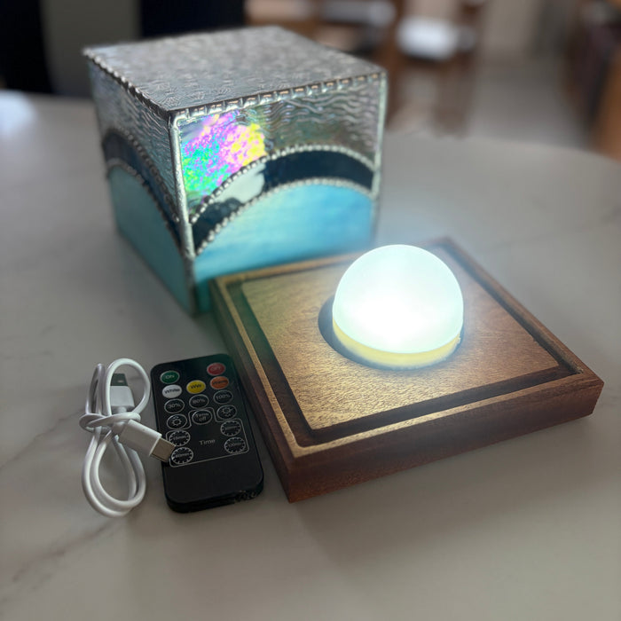 A square LED night light with a glowing white globe on a wooden base, shown with its textured metallic box packaging and a black remote control on a light marble tabletop.