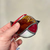 A hand holding a stained glass bird ornament with a red hanging string. The bird is made of amber, brown, red, white, and black glass sections outlined in dark metal, against a soft beige background.