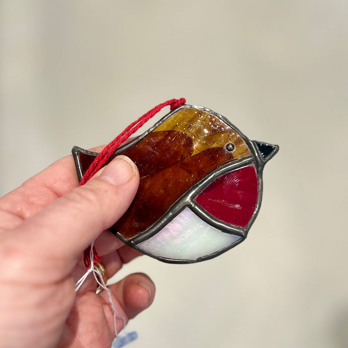 A hand holding a stained glass bird ornament with a red hanging string. The bird is made of amber, brown, red, white, and black glass sections outlined in dark metal, against a soft beige background.
