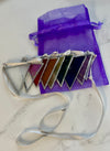 A set of seven triangular crystal pendant charms with silver borders and white ribbons, displayed next to a purple organza drawstring bag on a white marble surface.