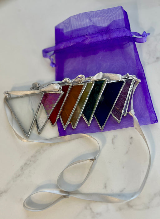 A set of seven triangular crystal pendant charms with silver borders and white ribbons, displayed next to a purple organza drawstring bag on a white marble surface.