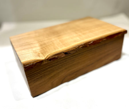 A rectangular wooden box with a live edge, featuring a light tan top and a darker brown side, photographed on a white surface with a soft-focus background.