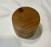 A small round wooden box with a lid, featuring a circular cutout near the top and visible wood grain, set against a white fabric background.