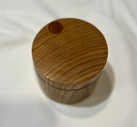 A small round wooden box with a lid, featuring a circular cutout near the top and visible wood grain, set against a white fabric background.