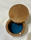 A round wooden keepsake box with its lid propped open, showing a bright blue velvet interior. The box is made of medium brown wood with visible grain patterns and small metal fasteners.