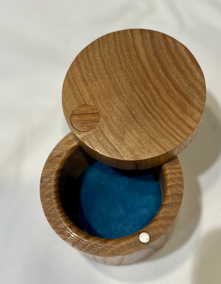 A round wooden keepsake box with its lid propped open, showing a bright blue velvet interior. The box is made of medium brown wood with visible grain patterns and small metal fasteners.