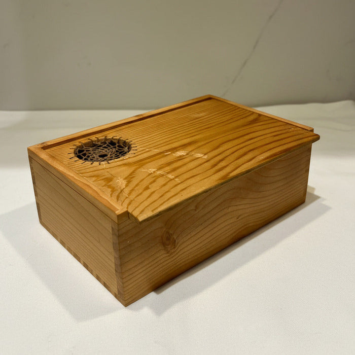 A rectangular wooden box with a sliding lid, made from light natural wood with visible grain. The lid features a circular cutout on the top-left corner containing a black spiderweb design.