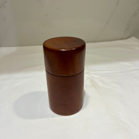A small cylindrical wooden container with a matching lid, made of dark reddish-brown wood, standing on a white fabric surface.