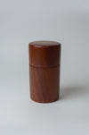 A cylindrical wooden container with a matching lid, made of medium brown wood, standing on a white surface with a blurred kitchen background.
