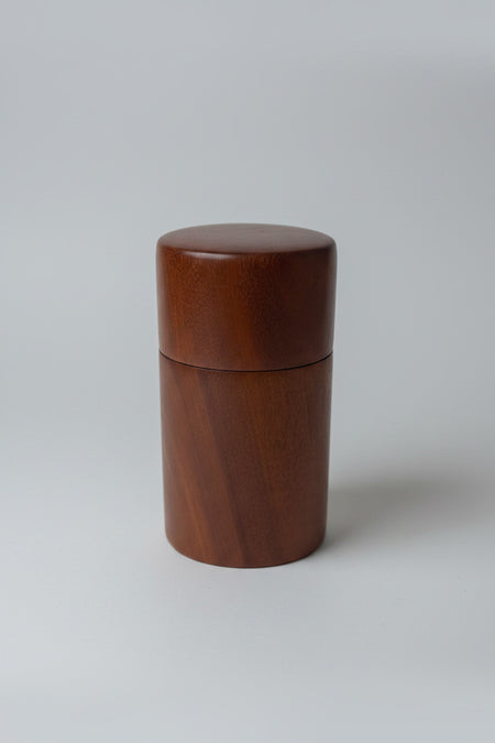 A cylindrical wooden container with a matching lid, made of medium brown wood, standing on a white surface with a blurred kitchen background.