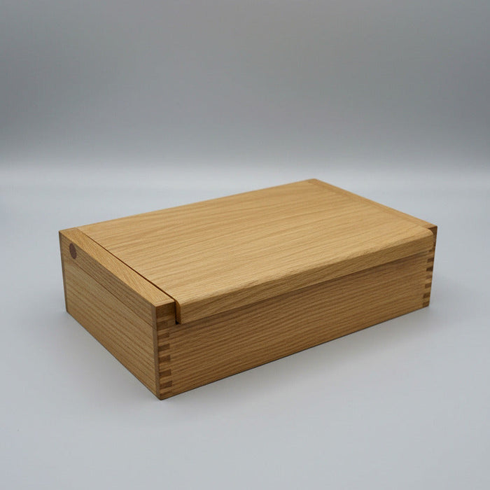 A rectangular light oak wood box with a sliding lid, resting on a white fabric surface with kitchen items blurred in the background.