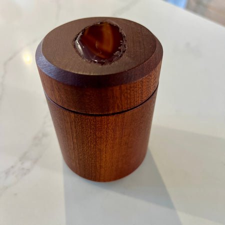 A small cylindrical wooden box with a lid, photographed on a white marble-like countertop. The top of the lid has a recessed oval area showing a brown polished agate piece.