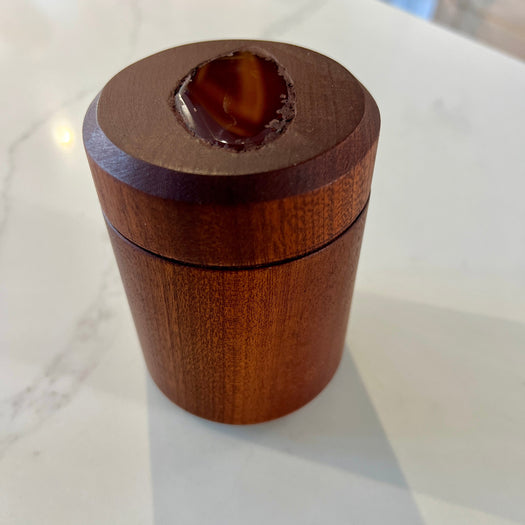 A small cylindrical wooden box with a lid, photographed on a white marble-like countertop. The top of the lid has a recessed oval area showing a brown polished agate piece.