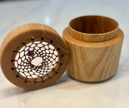A wooden keepsake box with its lid off, showing a natural wood grain exterior and a decorative inner tray with a web-like pattern and a small purple gemstone.