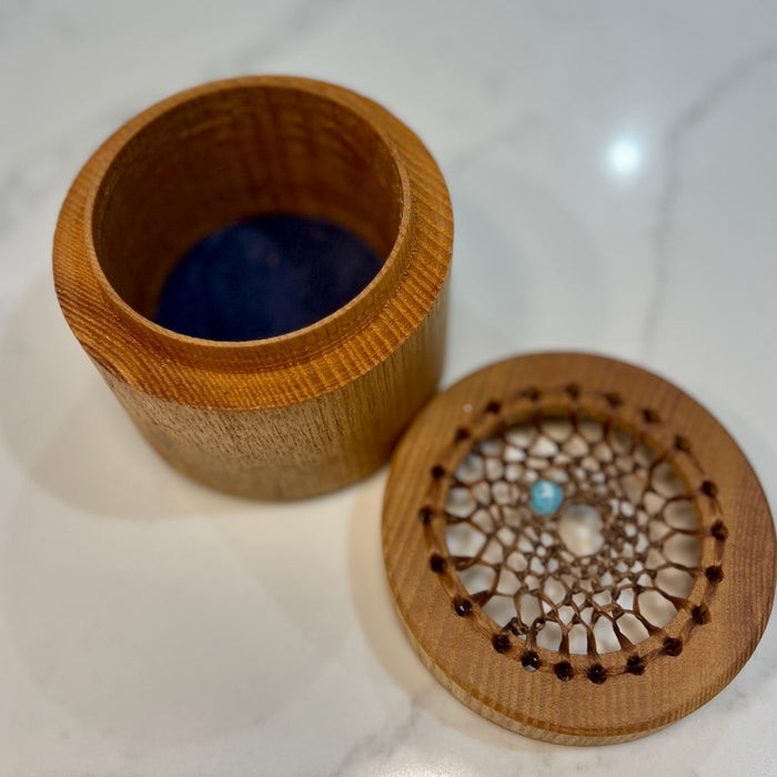 A round wooden box with its lid removed, placed on a light marble surface. The inside of the box is lined with dark blue material, and the lid features a laser-cut circular lattice design with a small light blue bead at the center.