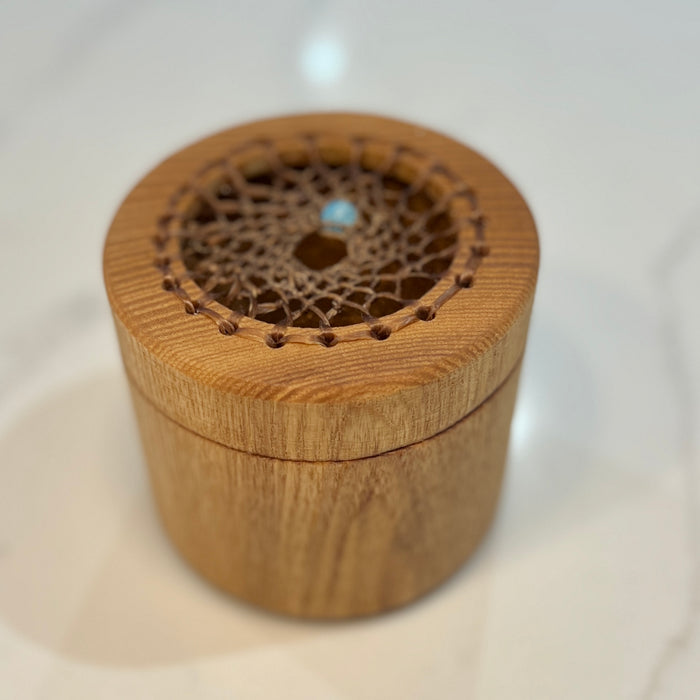 A small round wooden box with a lid featuring a decorative laser-cut lace pattern and a tiny light blue stone accent in the center, set on a light marble surface.