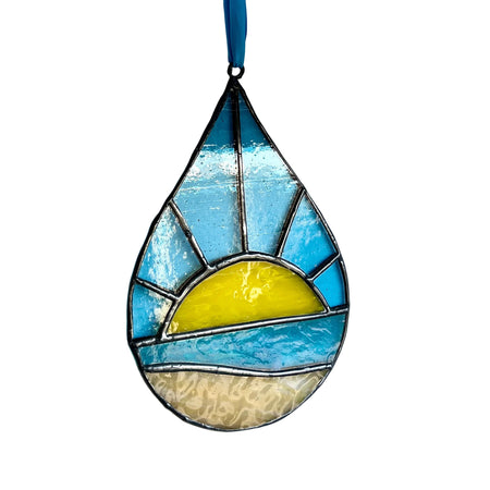 A teardrop-shaped stained glass ornament featuring a beach sunset design with blue sky, yellow sun, and beige sand, hanging against a plain white wall.