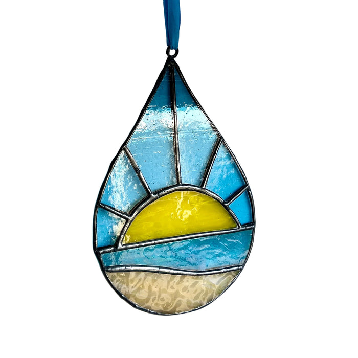 A teardrop-shaped stained glass ornament featuring a beach sunset design with blue sky, yellow sun, and beige sand, hanging against a plain white wall.
