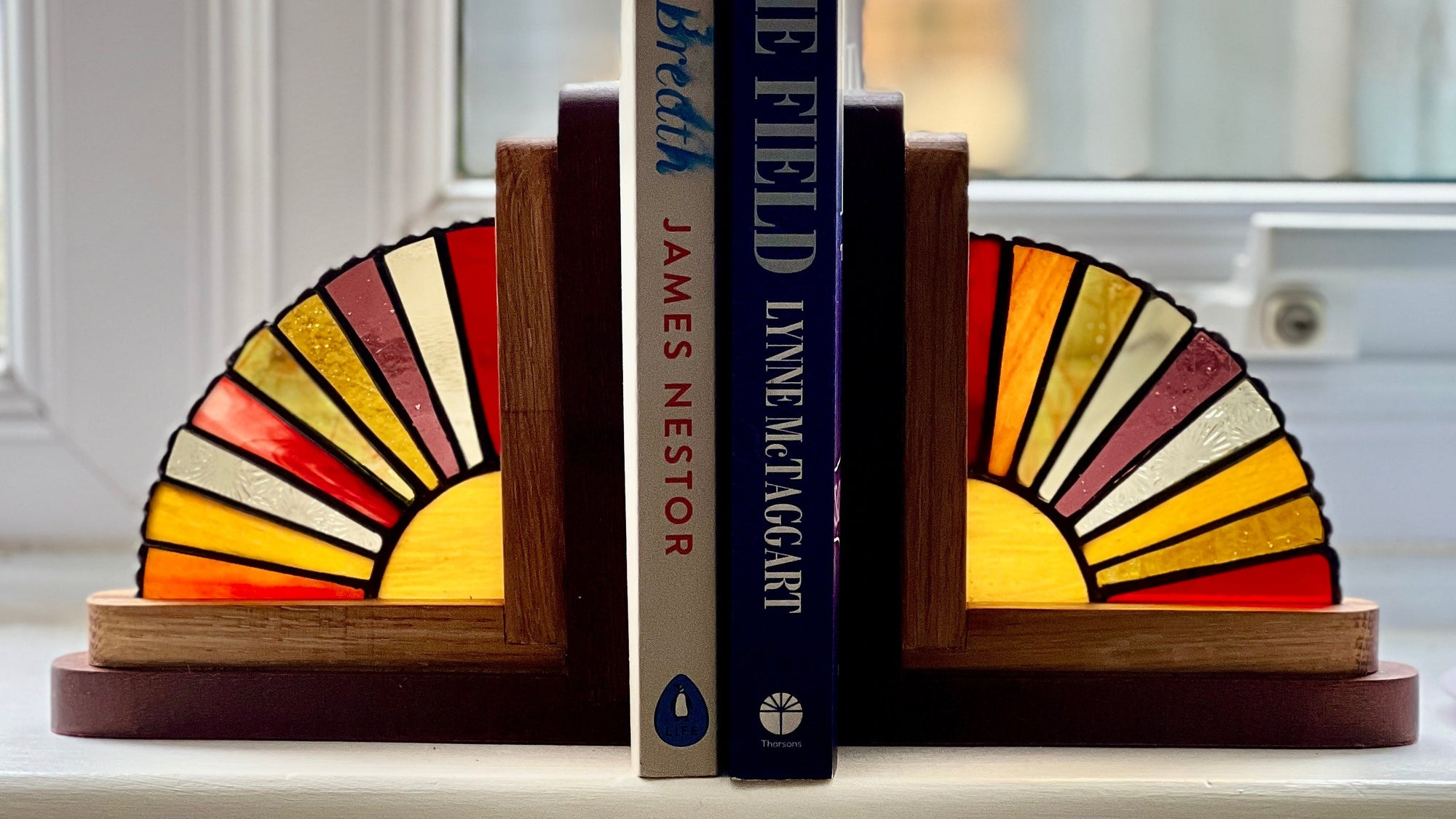 Two decorative wooden bookends with stained-glass-style fan designs in red, orange, yellow, cream, and brown, holding up three books on a windowsill.