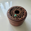 A round wooden box with a lid featuring a decorative woven bamboo pattern and a small green stone accent, photographed on a light surface.