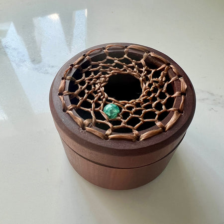 A round wooden box with a lid featuring a decorative woven bamboo pattern and a small green stone accent, photographed on a light surface.