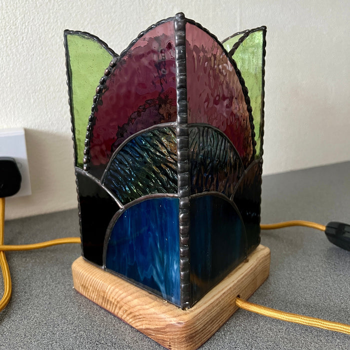 A stained-glass-style decorative pillar table lamp with scalloped shapes in green, burgundy, blue, and black, mounted on a light wood base on a gray tabletop against a beige wall.