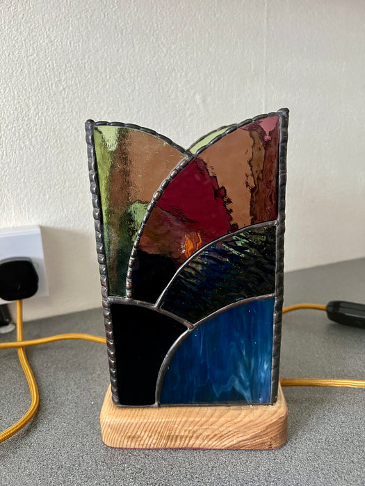 A stained glass table lamp with an abstract geometric design, featuring segments of blue, red, green, brown, and black glass, mounted on a light wood base.