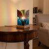 A stained glass table lamp with a geometric, fan-like Art Deco design, resting on a light wood base. The glass panels feature shades of burgundy red, olive green, dark blue, and black, separated by dark metal solder lines.